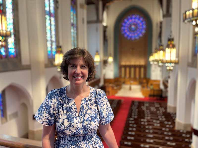 A person stands inside a church with stained glass windows and chandeliers, smiling. The interior features wooden pews and a rose window.