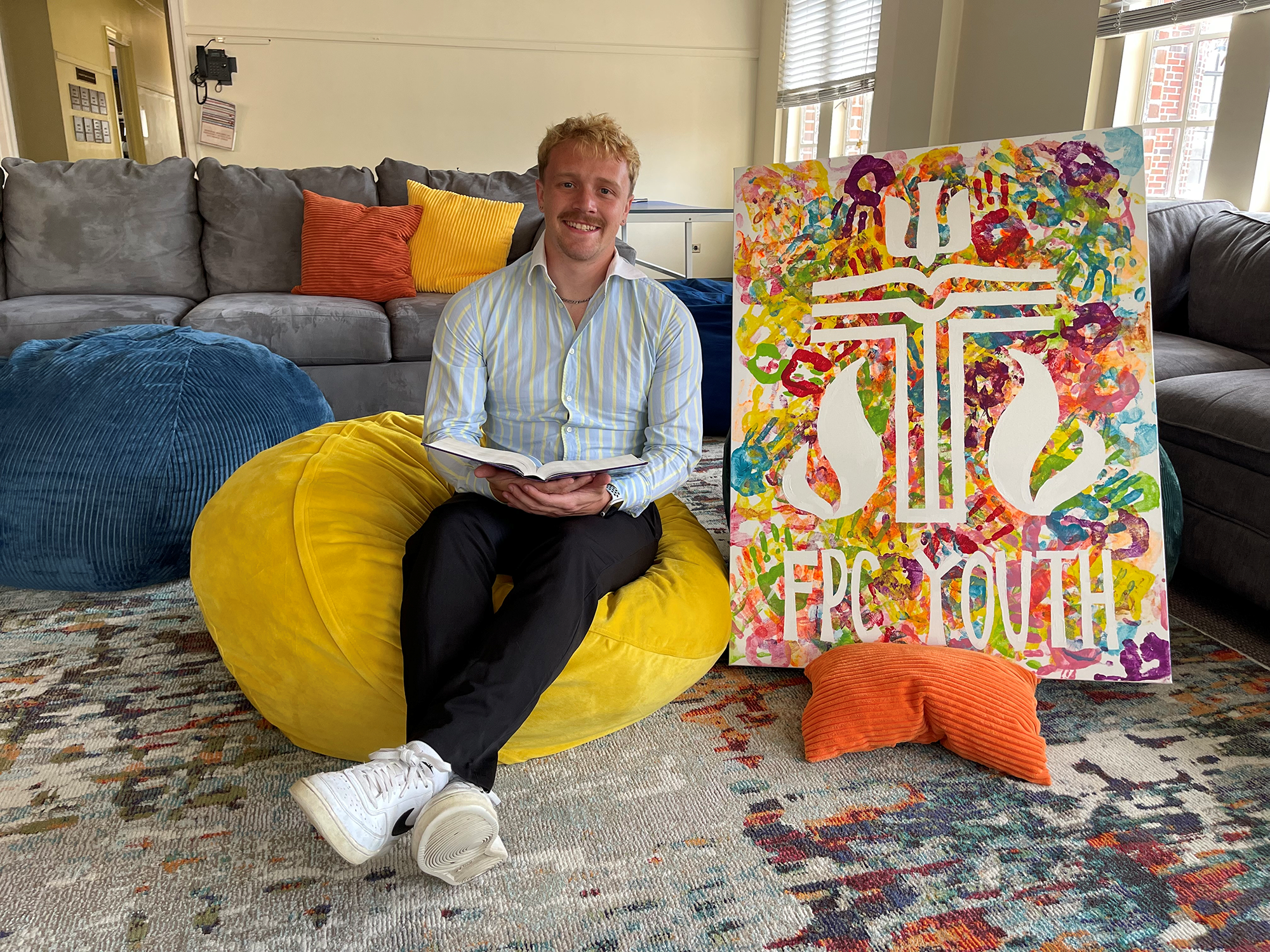 A person sits on a beanbag holding a book, next to a colorful artwork labeled “FPC YOUTH” in a cozy room.