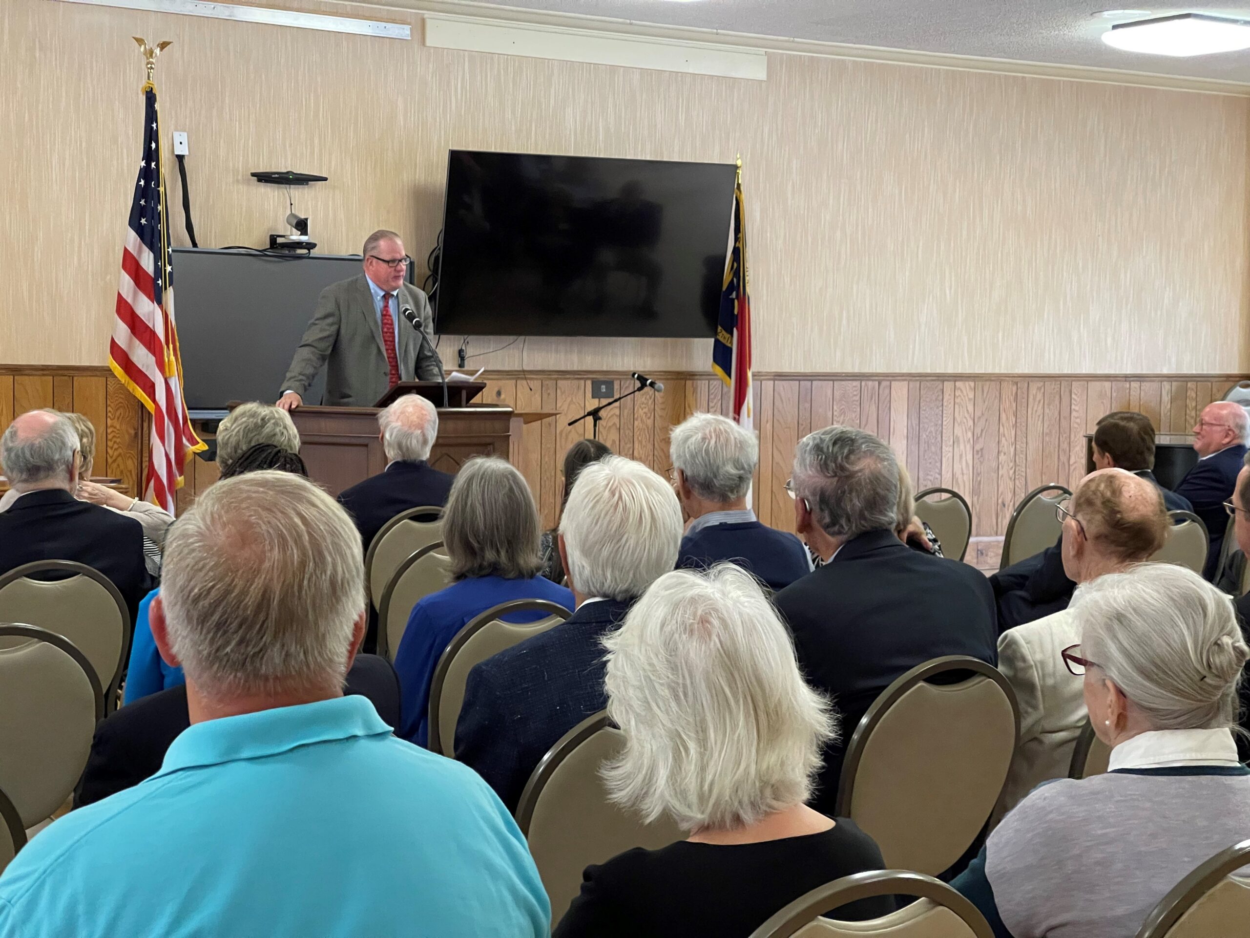 A person speaks at a podium to an audience in a hall, with flags and a television on the wall.