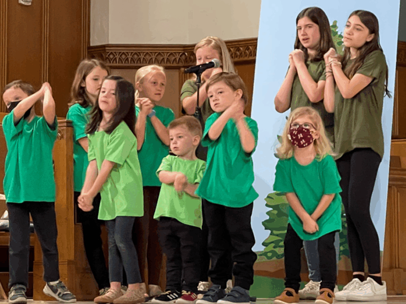 Children wearing green shirts sing onstage, with a person at the microphone. A nature-themed backdrop is visible behind them.