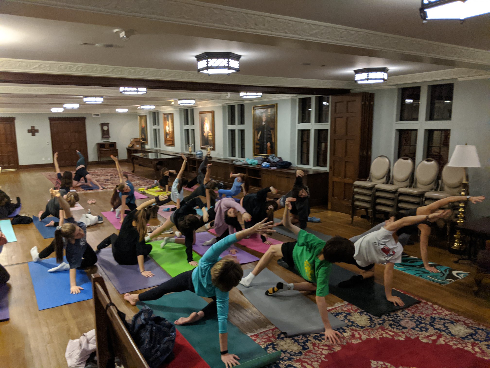 People and children practicing yoga on colorful mats in a spacious, carpeted room with wooden furniture and painted portraits on the walls.