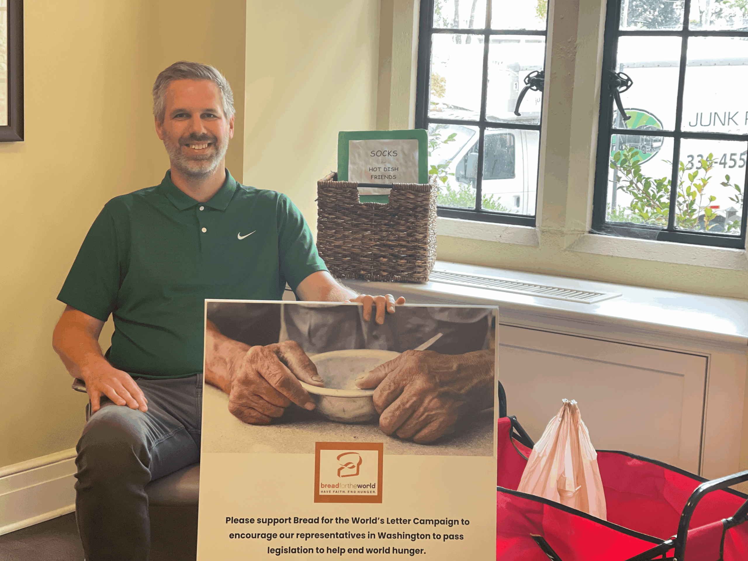 A person in a green shirt sits indoors, holding a sign about ending world hunger. A window and woven basket are behind.