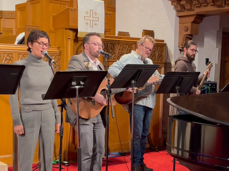 Four people perform with guitars and microphones in a church setting, standing near wooden paneling and a grand piano.