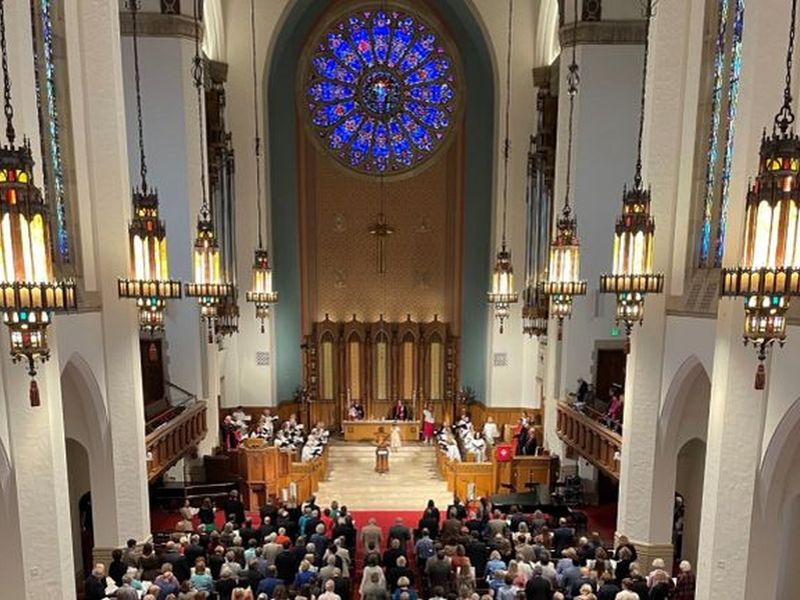 Large church interior with stained glass rose window, ornate chandeliers, and people gathered for a service. The altar area is prominent and decorated.